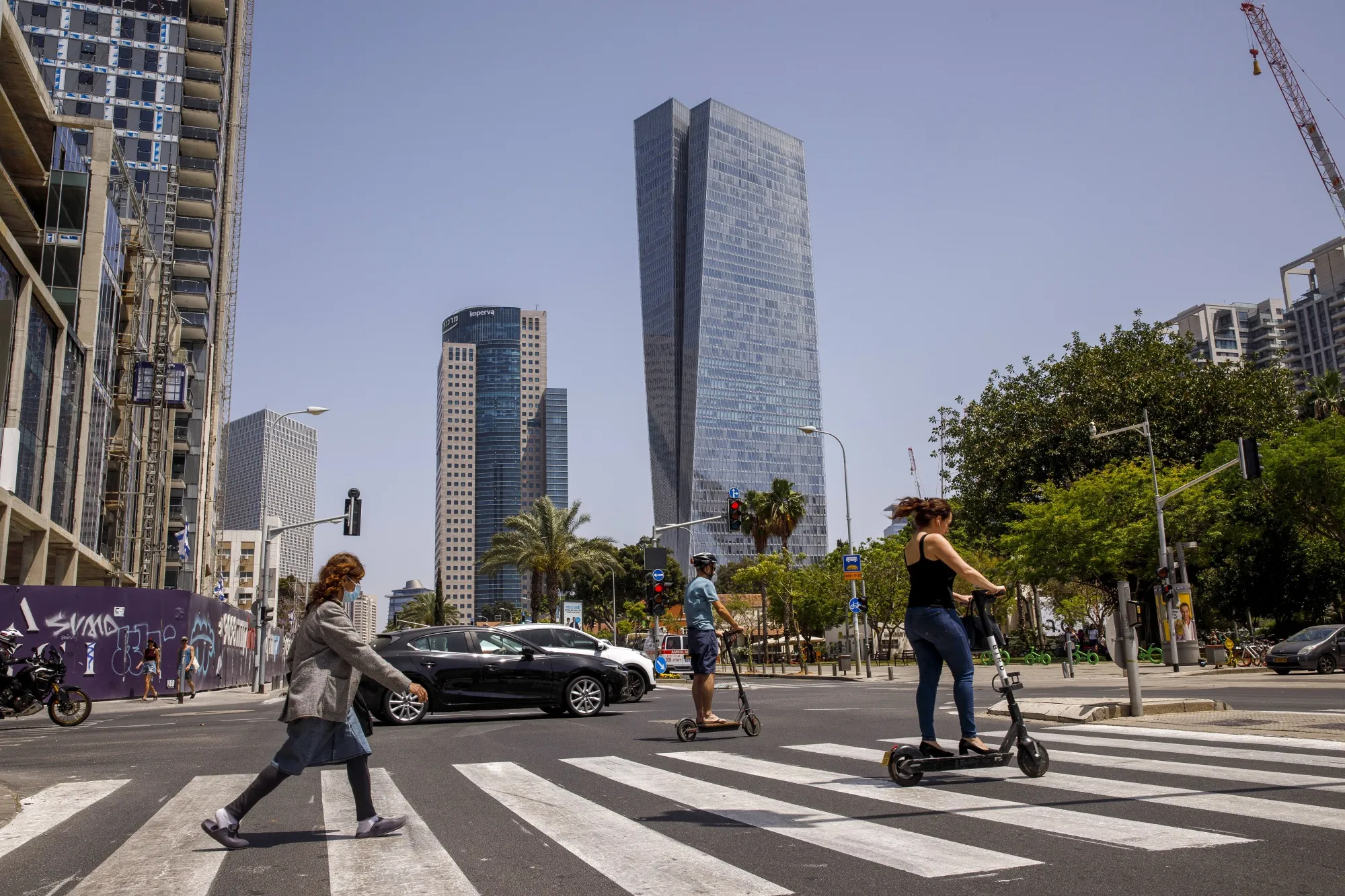 Commuters cross a street with the Azrieli Tower skyscraper in the background on April 30, 2021. An influx of tech workers has brought more e-scooters to Tel Aviv’s streets. 