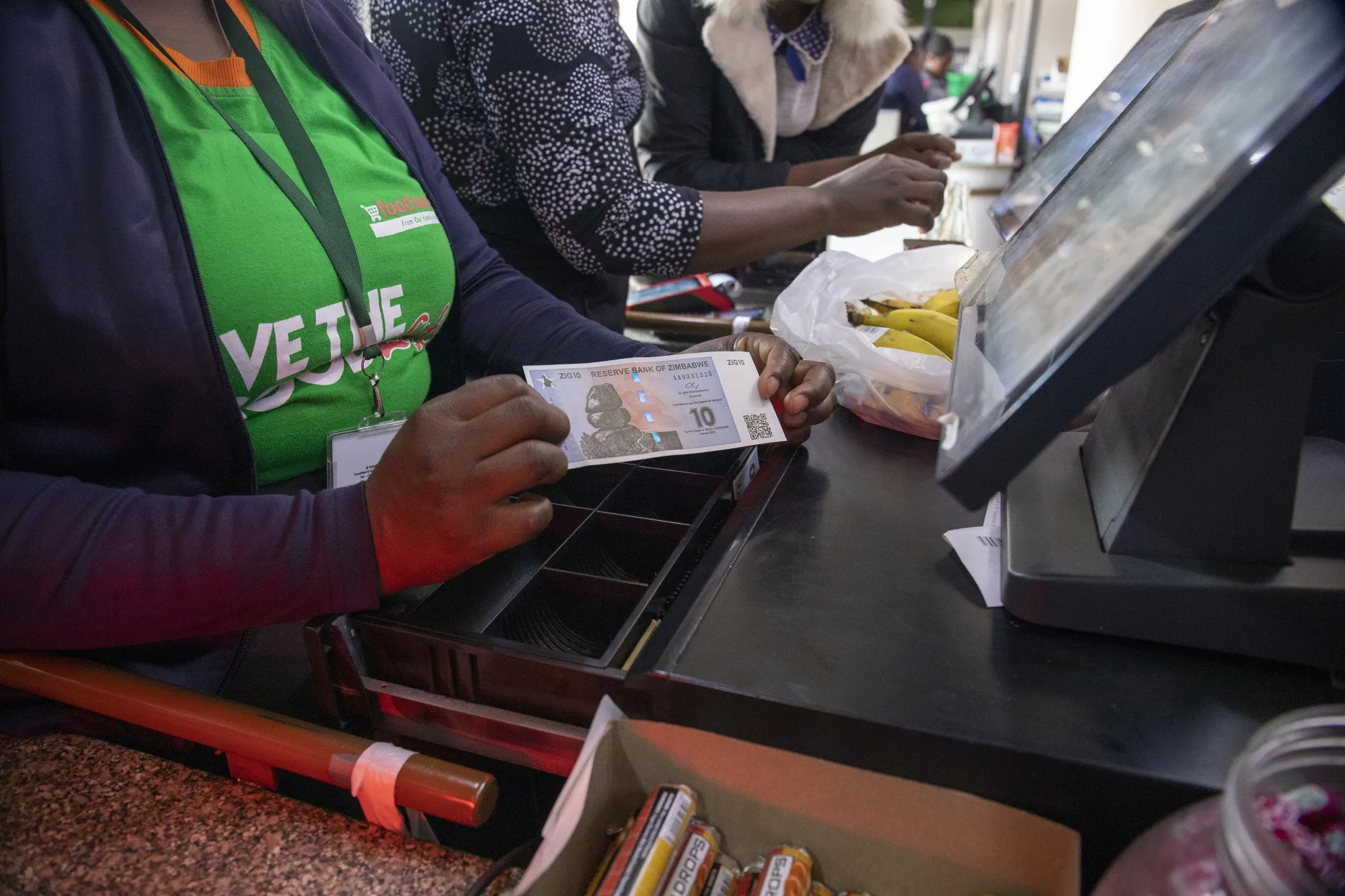 A supermarket till operator displays a 10 ZiG banknote.