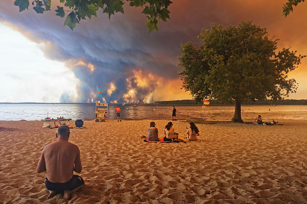 TOPSHOT - Beach goers rest on the beach of Moutchic on the shore of Lake Lacanau (Etang de Lacanau) in Lacanau , southwestern France on July 24, 2026, as clouds of smoke rise into the sky from the Gironde wildfire. The fire raging in the northern part of the Arcachon Basin in Gironde, southwest France, has swept through more than 3,400 hectares, according to the latest figures released by the prefecture. More than 20,000 residents and tourists have been evacuated from this part already devastated by flames in 2022. Scientists have long warned that climate change caused by human use of fossil fuels will make periods of drought more intense and longer-lasting, creating the ideal conditions for wildfires. (Photo by Maximilien LAMY / AFP via Getty Images) Photographer: MAXIMILIEN LAMY/AFP