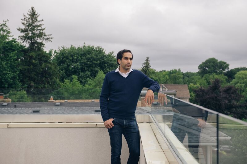 Man in sweater and jeans posing on a roofdek of a home with a gray sky and trees in the background