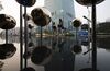 Two young women walk by a sculpture installation of large metallic spheres on poles in a shallow pool with scooters and taxis in the background, across the street from the Taipei 101 building