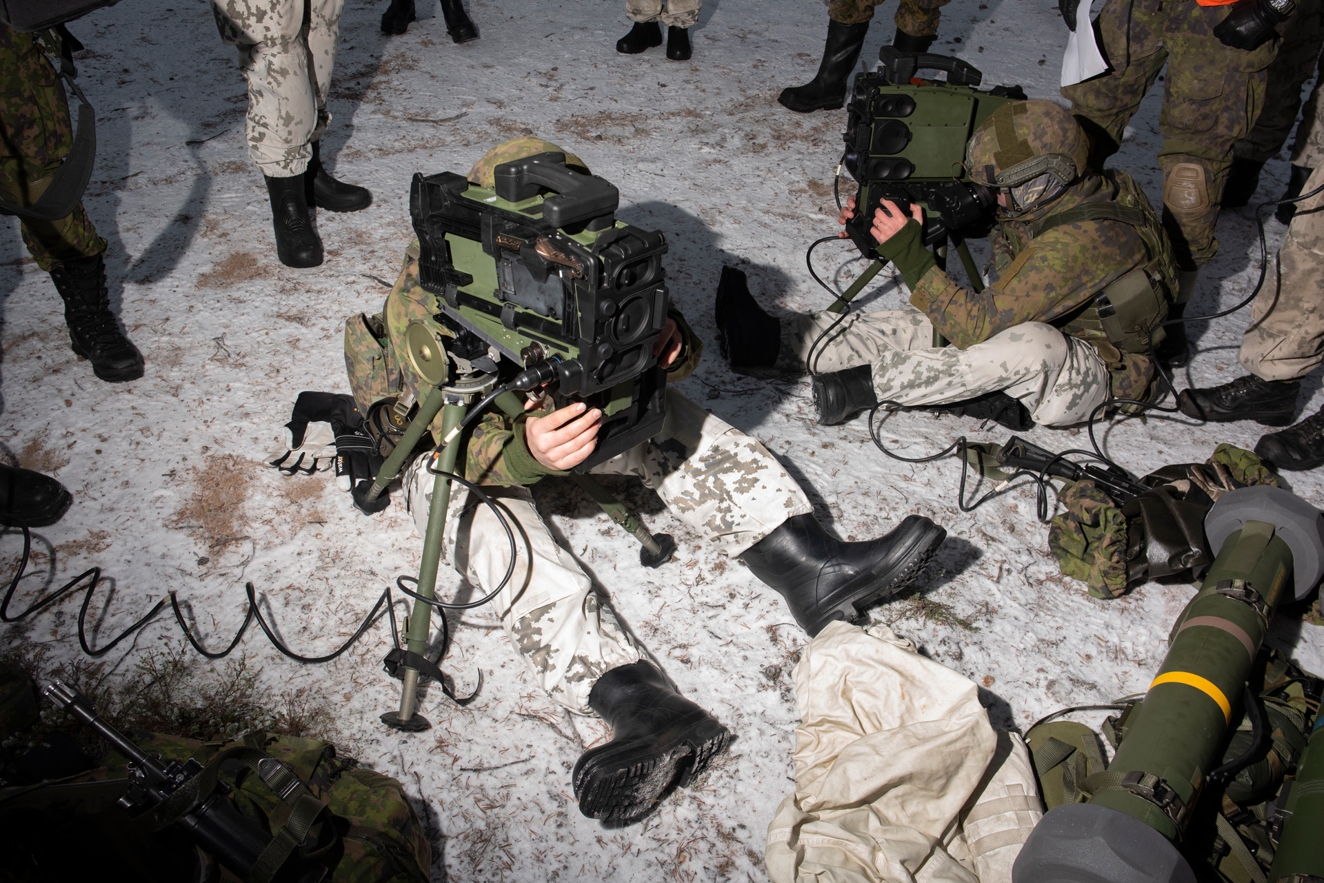 Two Finnish soldiers sit on snow covered ground looking into the viewfinders of bulky sighting equipment that resembles TV cameras, mounted on small tripods. They look in different directions, their faces obscured by the equipment. They wear white camouflage on their legs and green camouflage uniforms on their upper body. The legs of other soldiers in a mix of white and green camouflage can be seen as they stand around the edge of the frame. The bulky green tube of an anti-tank weapon can be seen to the right.