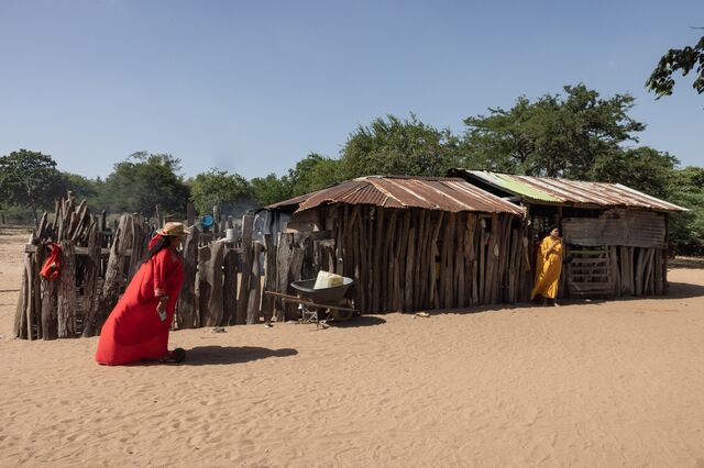 Remedios Sierra, left, in a red dress walks in front of a home while a resident in a yellow dress stands outside holding a beverage in a cup.