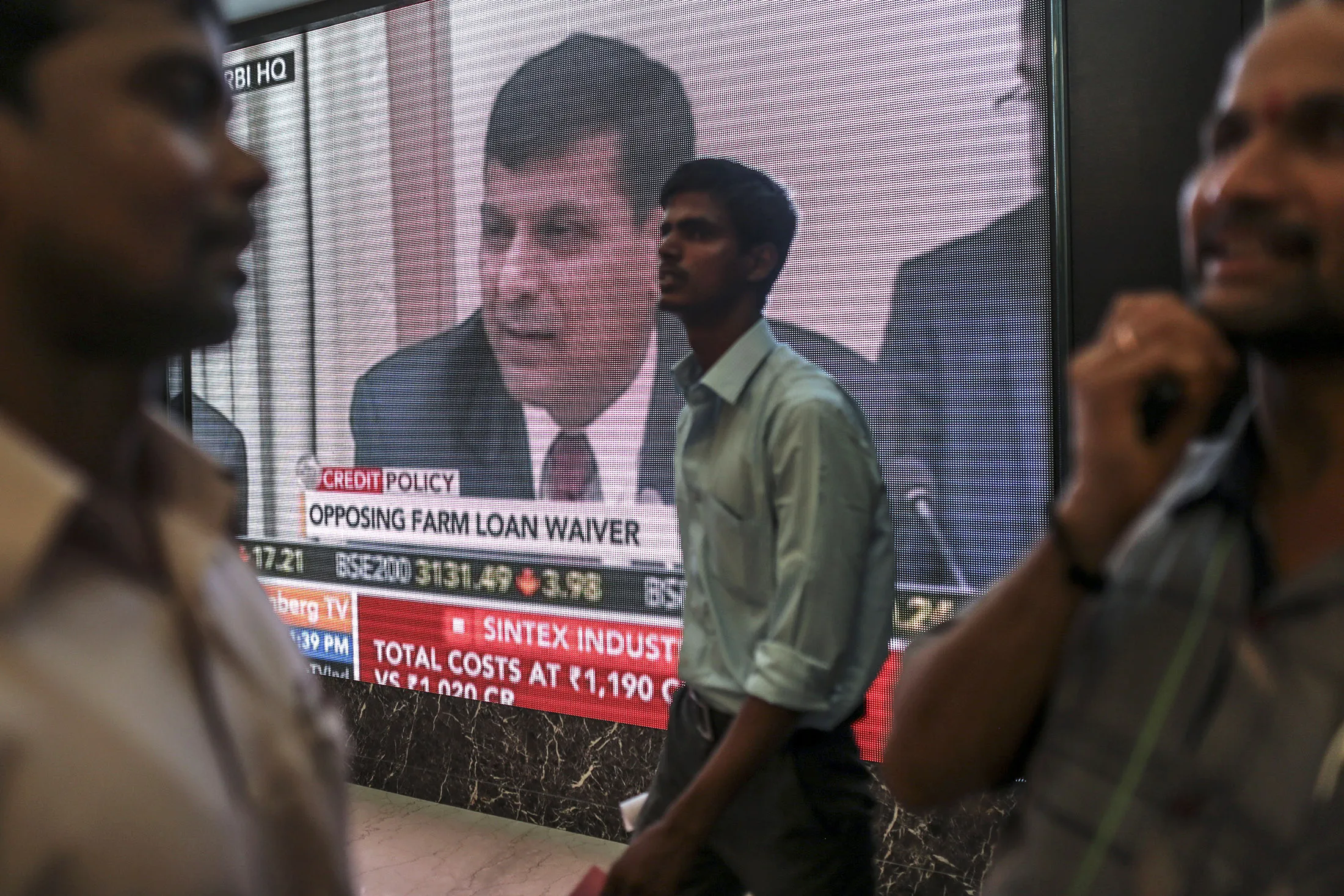People walk past an electronic screen showing television coverage of Raghuram Rajan, governor of the Reserve Bank of India (RBI), speaking during a news conference at the central bank's headquarters, in the lobby of the Bombay Stock Exchange (BSE) in Mumbai.
