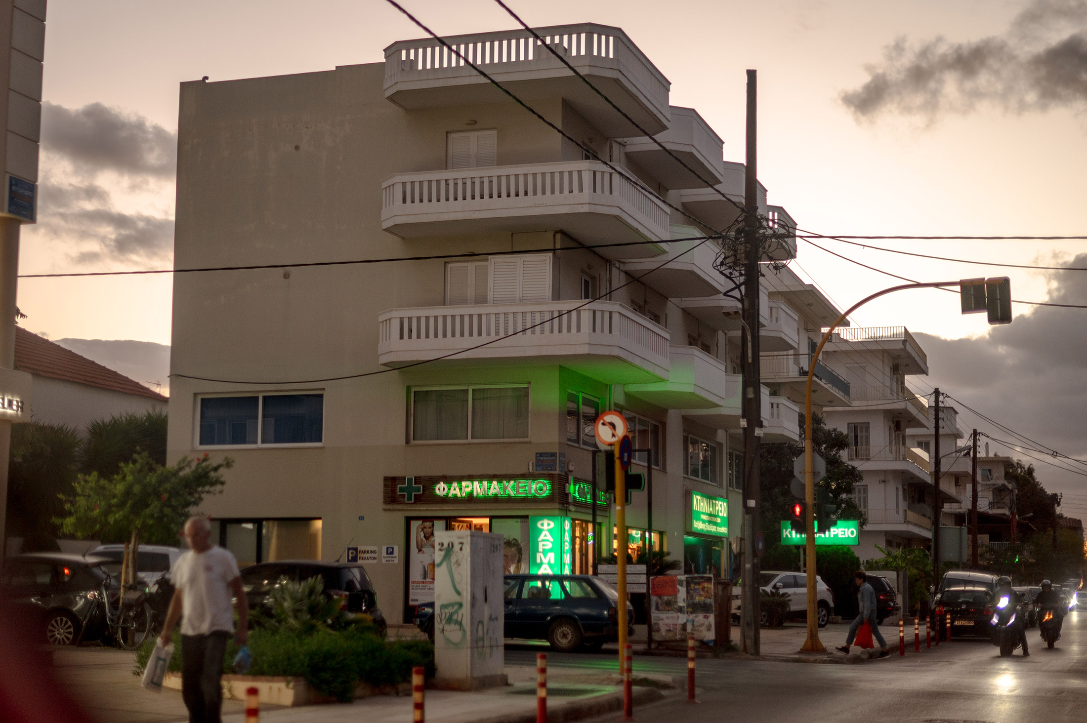 A three story building at dusk with balconies and lit green pharmacy sign. Pedestrians, cars and motorbikes are in the streets and sidewalks. 