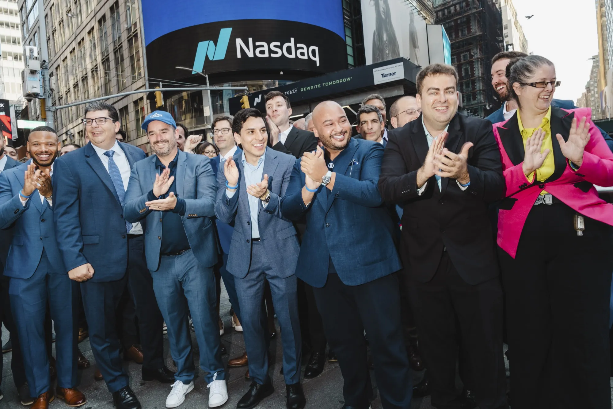 A crowd of mostly men in blue or black suits smiles and applauds. The Nasdaq logo is visible on a building in the background.