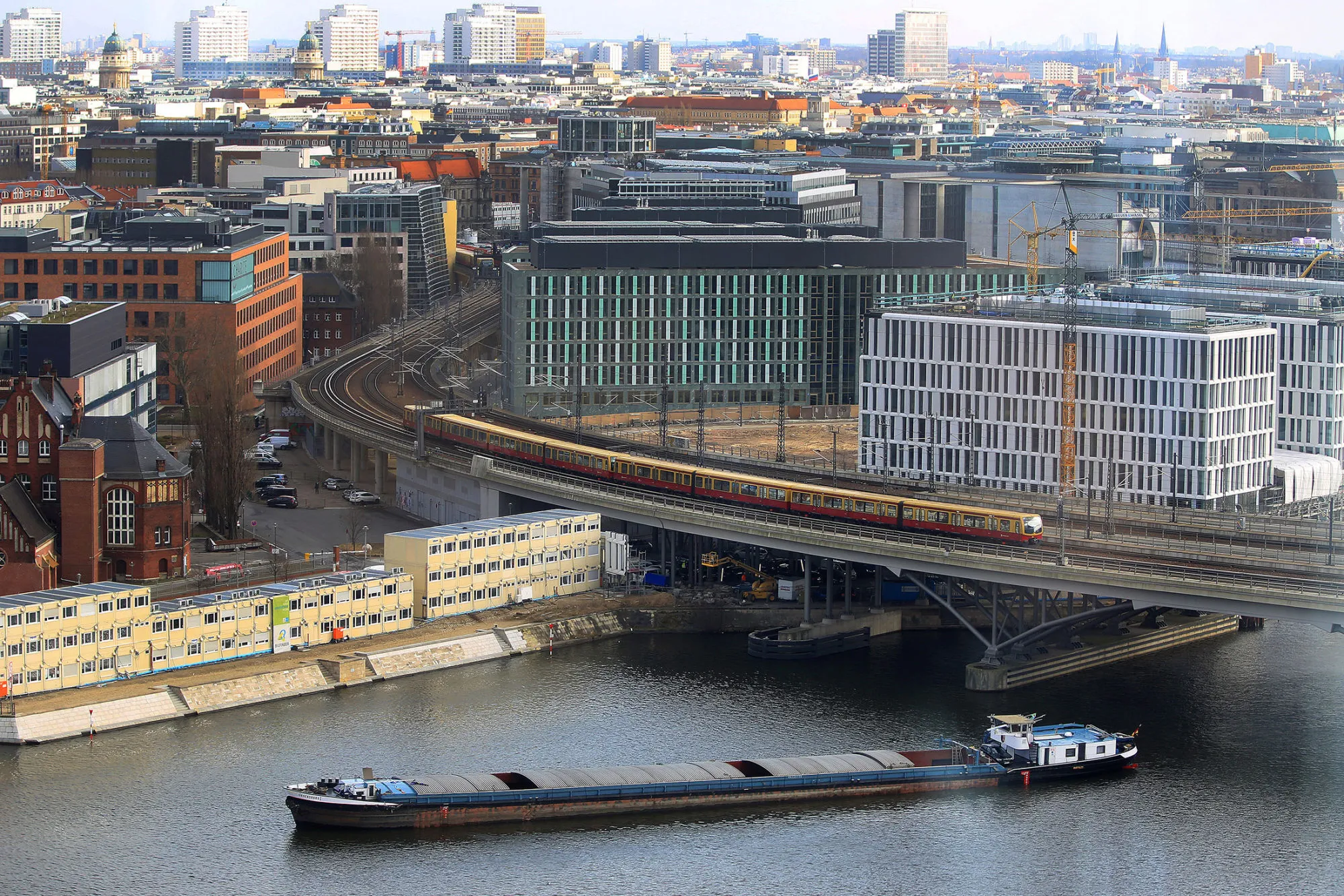 An S-Bahn passenger train travels along a bridge on route to Berlin Central railway station as construction cranes stand on the city skyline and a cargo barge sails on the river Spree in Berlin.
