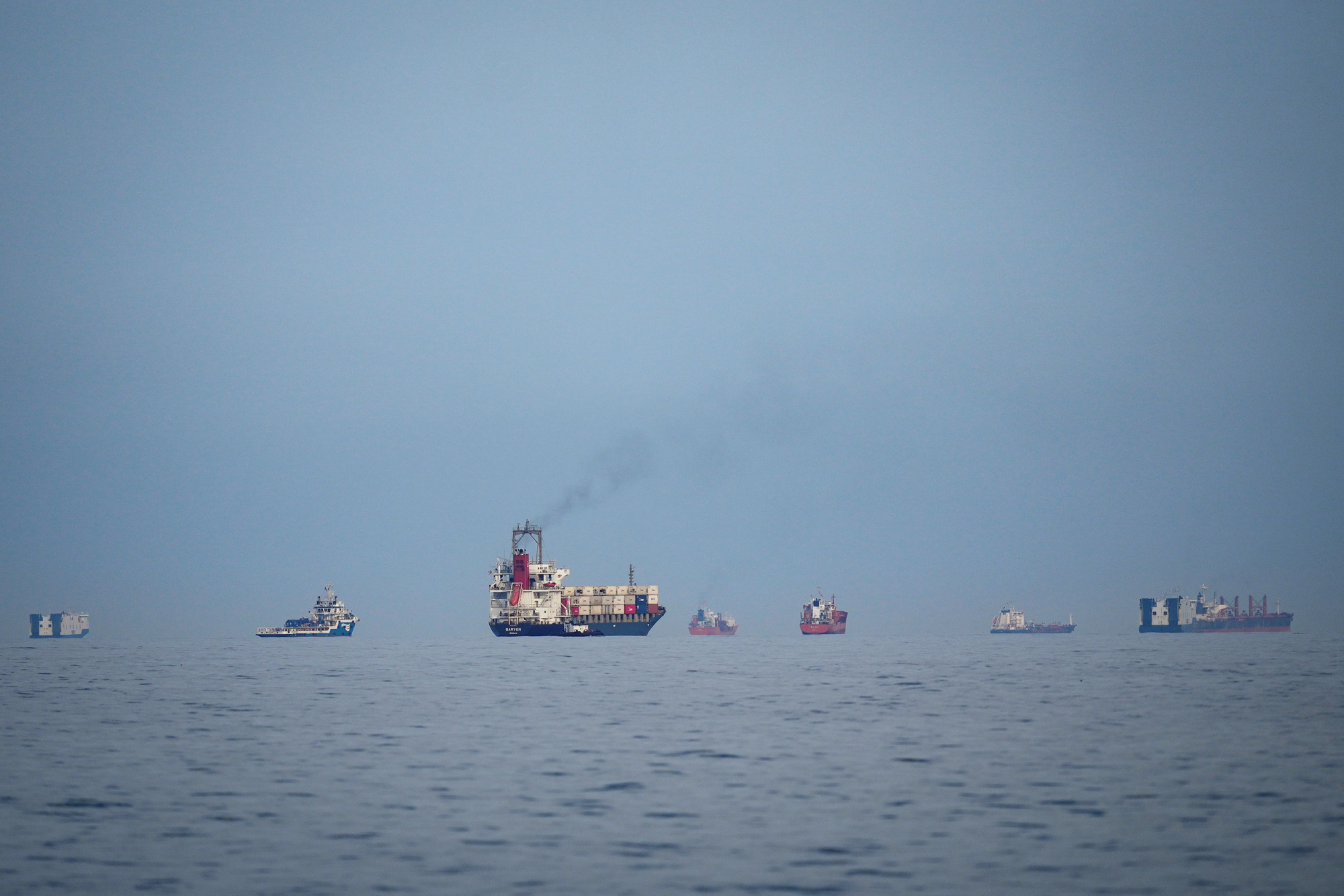 A wide-angle photograph taken from the water shows a diverse group of multiple commercial vessels, including a container ship emitting a plume of dark smoke, anchored along a hazy horizon under an overcast sky.