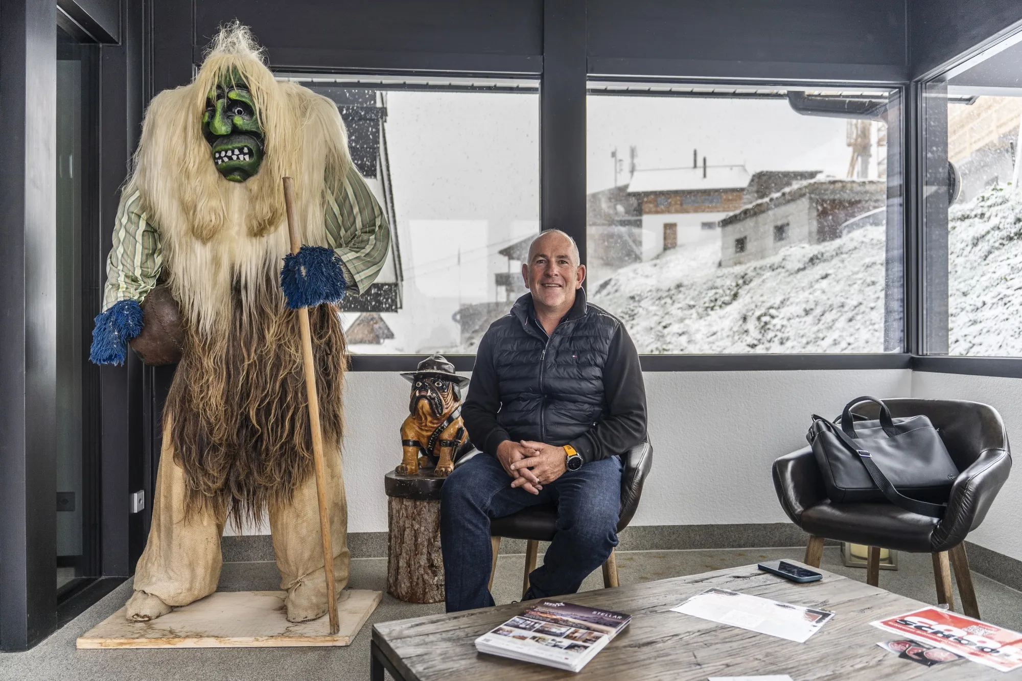 A man sits in a chair. Through windows behind him is a snow-covered town. Next to him is a large creature with fur, holidng a stick, wearing a mask.