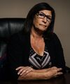 Carol Davis sits at a desk with her arms crossed and a serious expression. She has long, dark hair and is wearing glasses and a black and white top.