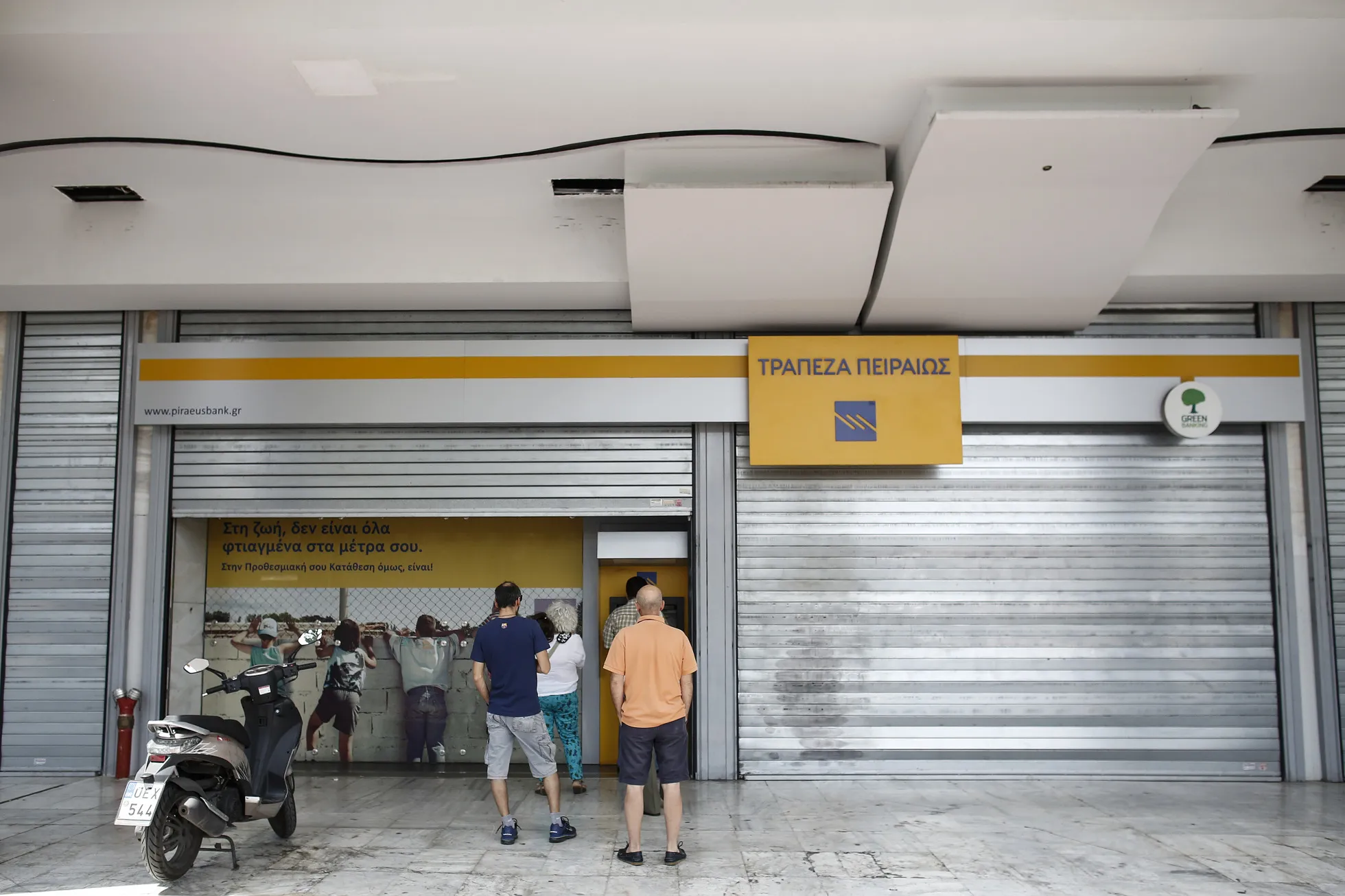 Customers queue to use an ATM&nbsp;outside a shuttered Piraeus Bank SA bank branch in Athens, Greece.