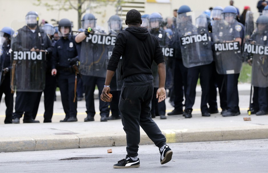 A demonstrator walks past police with a brick as they respond to thrown objects, Monday, April 27, 2015, after the funeral of Freddie Gray in Baltimore.