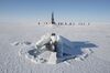 The sail and parts of the stern of the USS Connecticut, a US Navy attack submarine, break through sea ice off the north coast of Alaska with crew in red or dark-colored uniforms standing in the distance against a backdrop of a cloudless, blue sky.