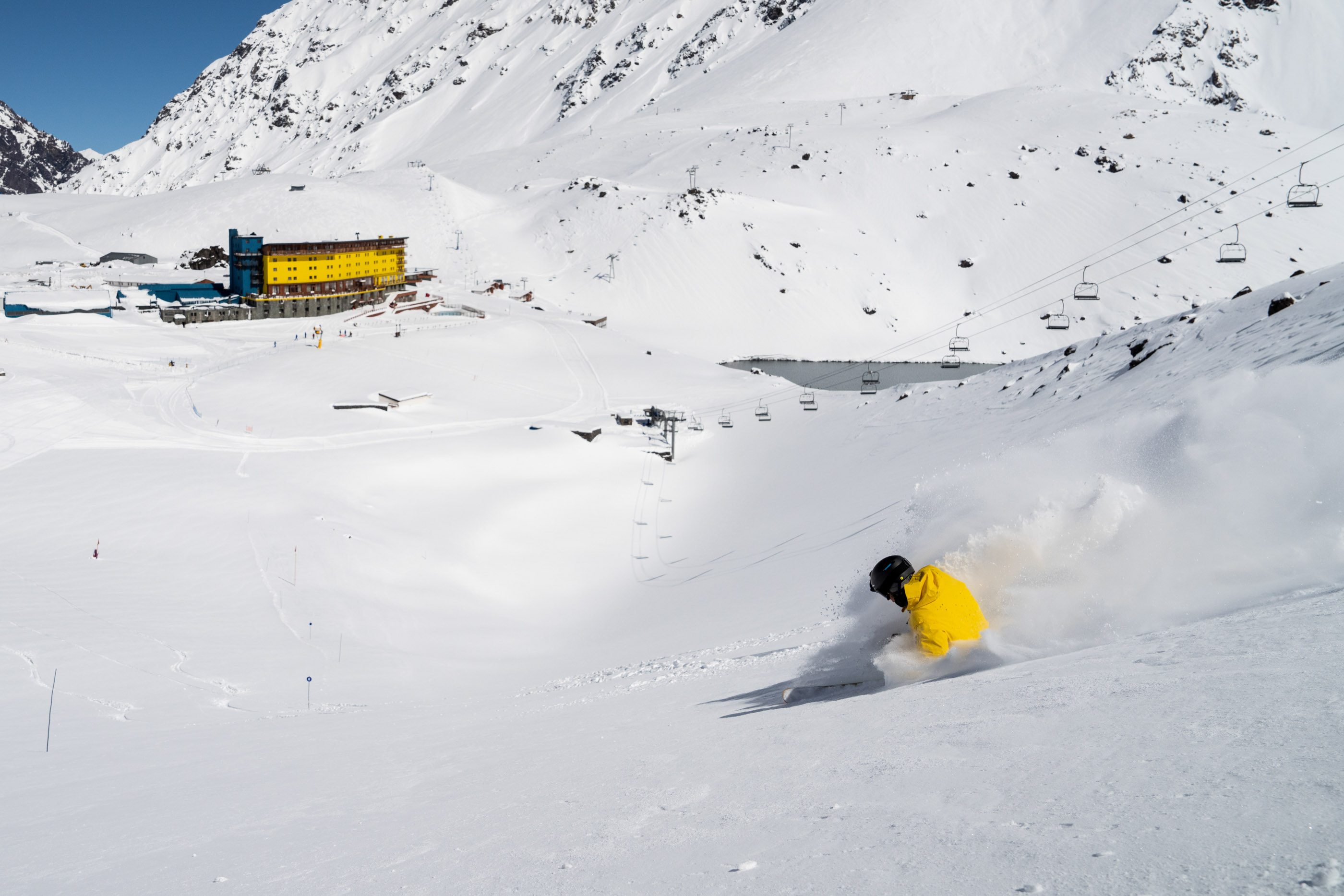 A skier makes their way down an otherwise empty slope, with the yellow facade of the Portillo Hotel in the distance.