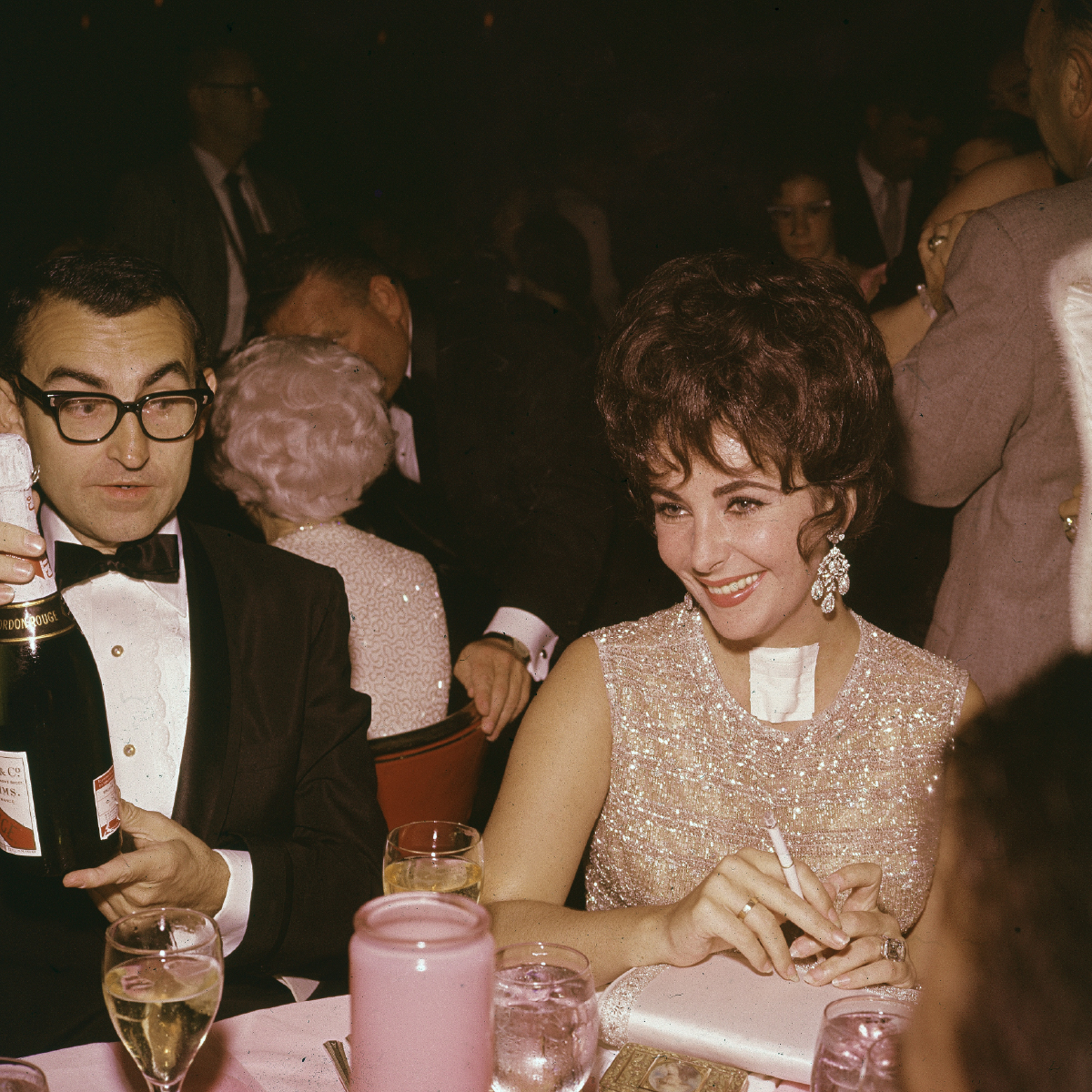 Elizabeth Taylor smiles with a cigarette in hand. She is formally dressed in a sparkly dress, wearing a throat bandage, as a man in a tailored tuxedo raises a bottle of champagne at the Coconut Grove nightclub in California, 1961