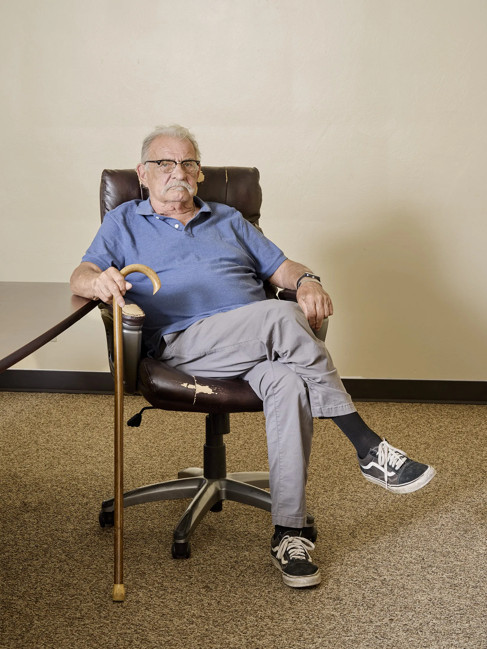 Shoen sits in a tattered brown leather office chair, with his legs cross and facing the camera. He is wearing black glasses and black Vans sneakers, has white hair and a mustache, and is holding a cane.