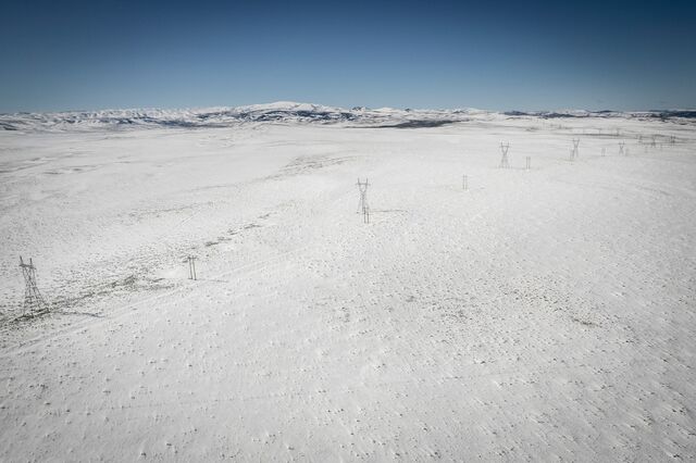 A photo of power lines taken by a drone from above otherwise undeveloped flat land, with snow on the ground and mountains in the distance.