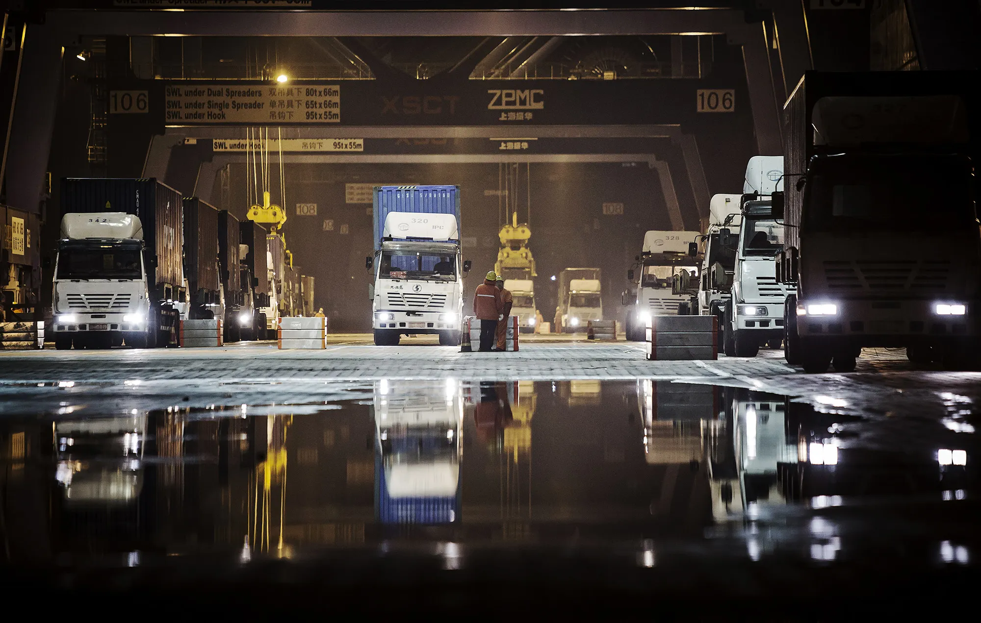 Trucks&nbsp;wait in line to unload their containers onto a&nbsp;container ship docked at the the Xiamen Songyu Container Terminal at night in Xiamen, China.