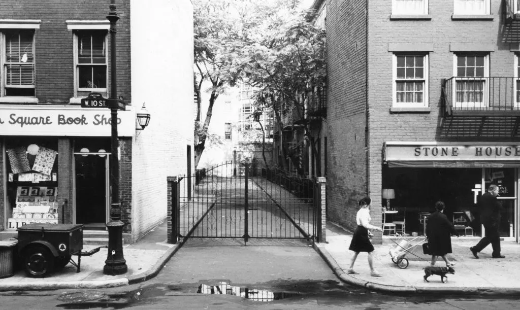 An archival black and white image of a woman walking her dog and other pedestrians on West 10th street infron of a gated cul-de-sac in Greenwich Village.