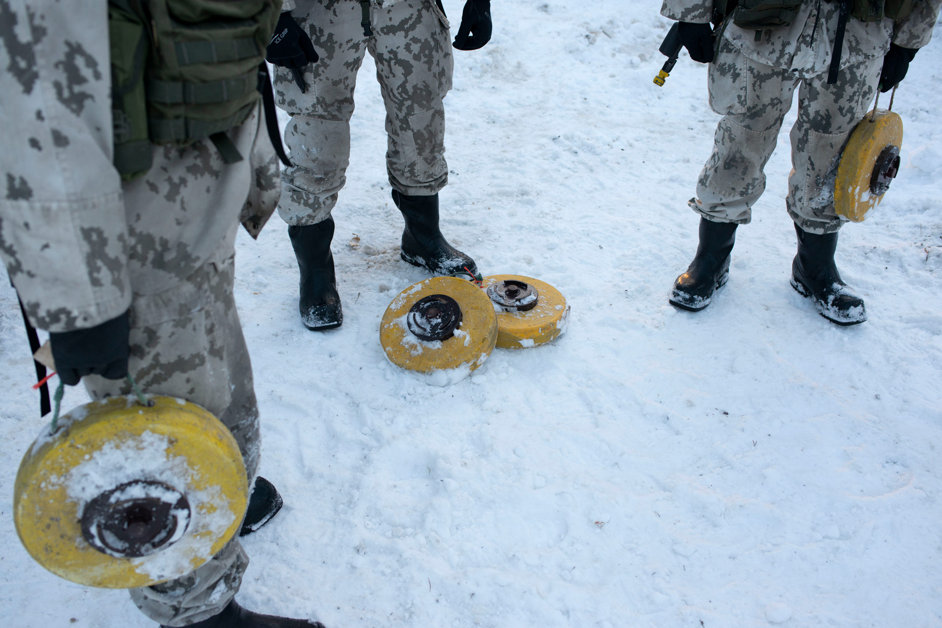 The legs of three Finnish soldiers are framed with the camera pointed down toward the snow-covered ground. They all wear white camouflage uniforms and black boots. Two of them grip handles attached to round, yellow anti-tank mines that resemble a discus used in athletics. Two more mines lie at the feet of the third soldier.