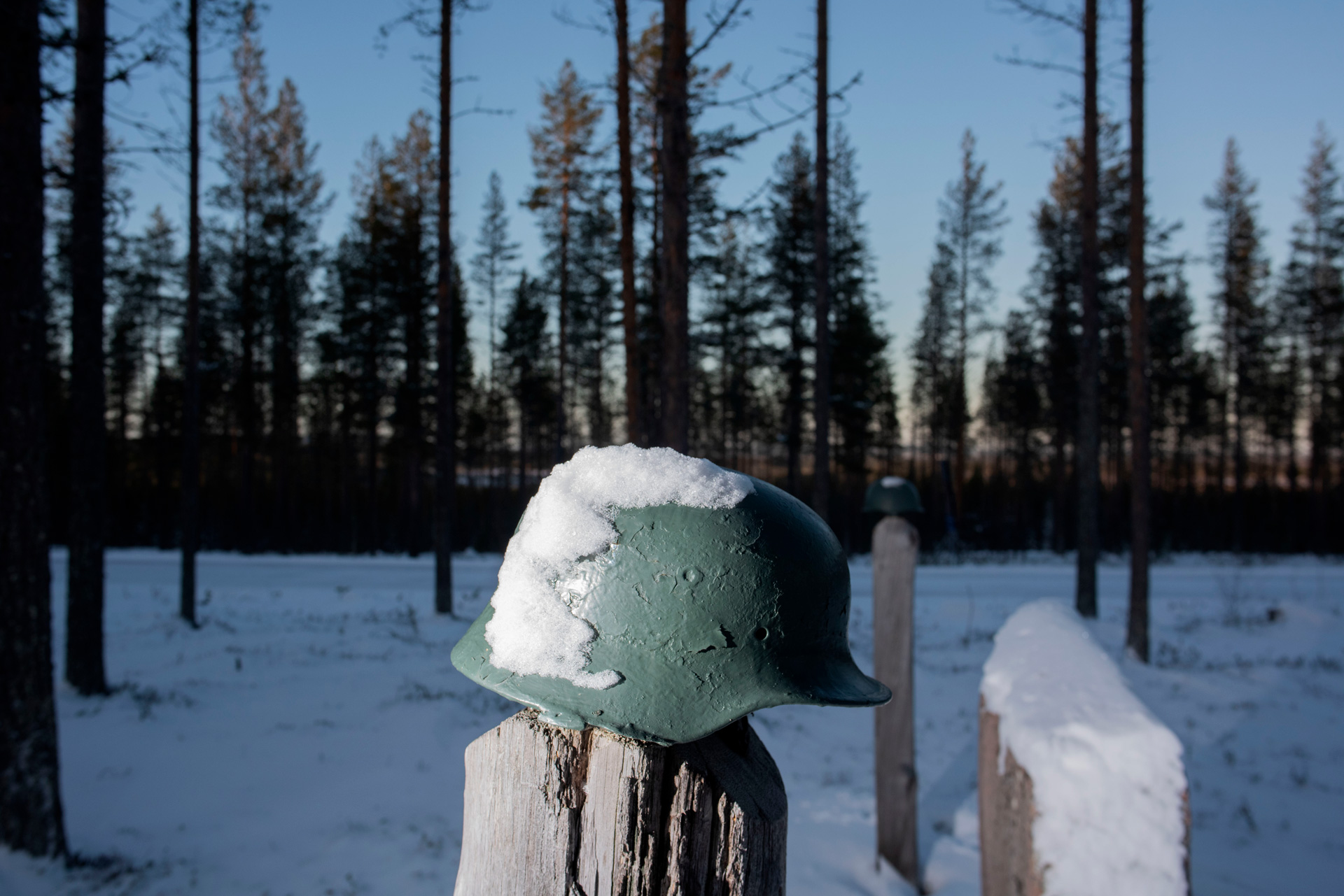 An old-fashioned, green military helmet sits atop a rough wooden post in the foreground. It is partly covered with snow. The post sits in a snowy Finland landscape against a backdrop of trees and a clear blue sky at twilight.