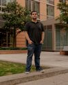 A man stands on a concrete curb in front of a beige brick apartment building. He has short, dark brown hair and is wearing jeans and a black polo shirt, where a black tattoo peeks out from one of the sleeves.