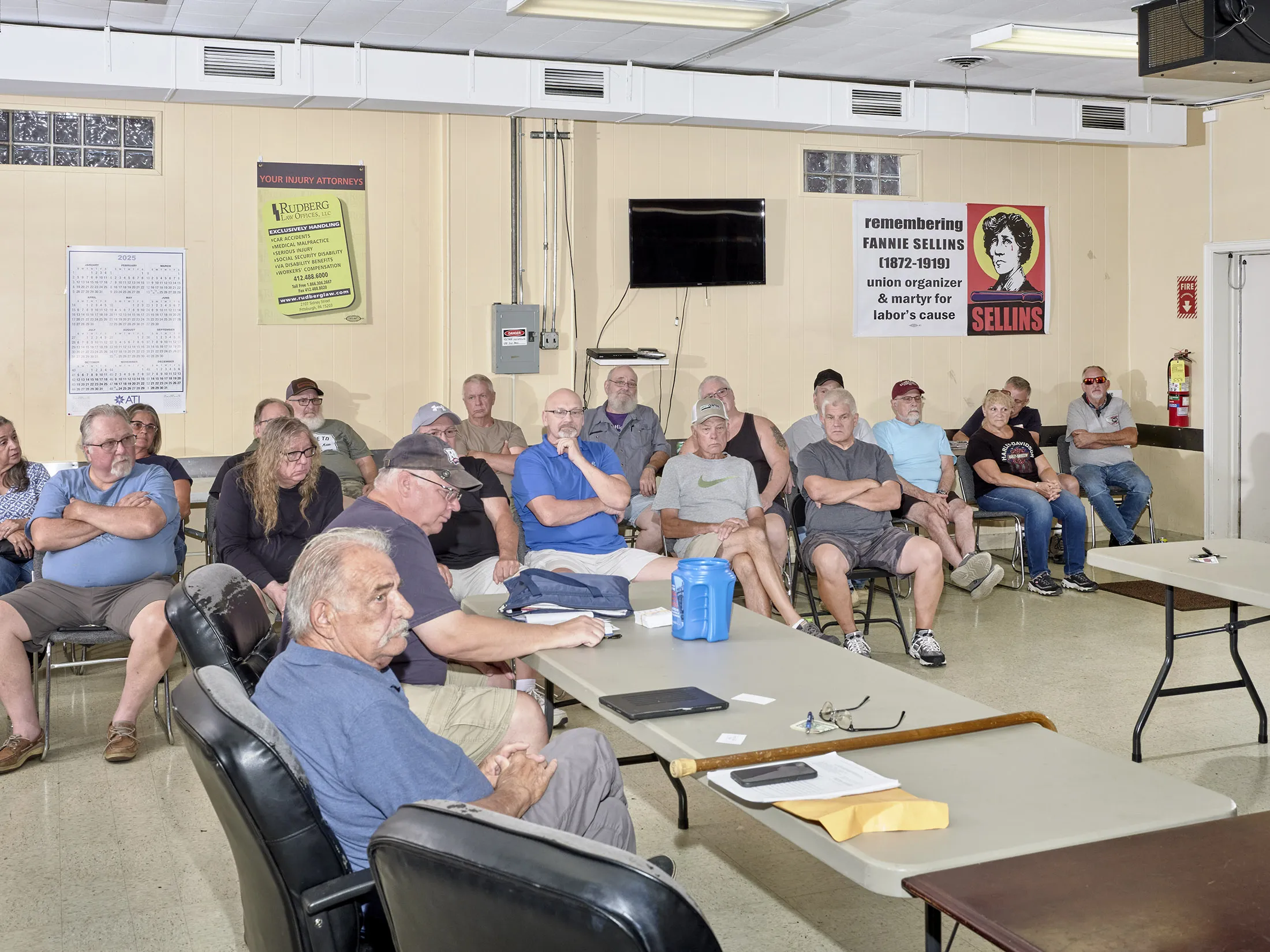 The group of retired steelworkers sit at chairs and folding tables, their gazes focused on something out of the frame of the image. Behind them is a calendar, a poster for an injury attorney and a memorial poster for the labor activist Fannie Sellins.