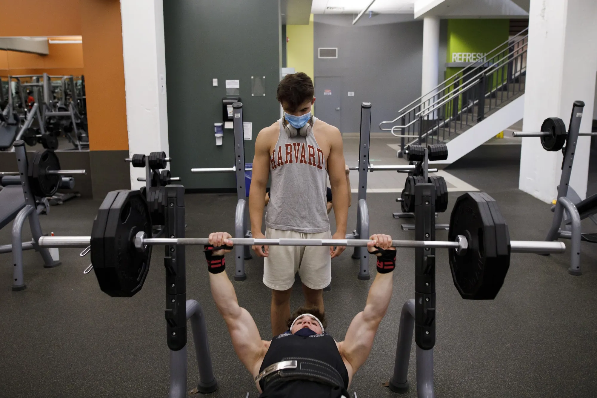 A person wearing a protective mask spots a person lifting weights at a gym.