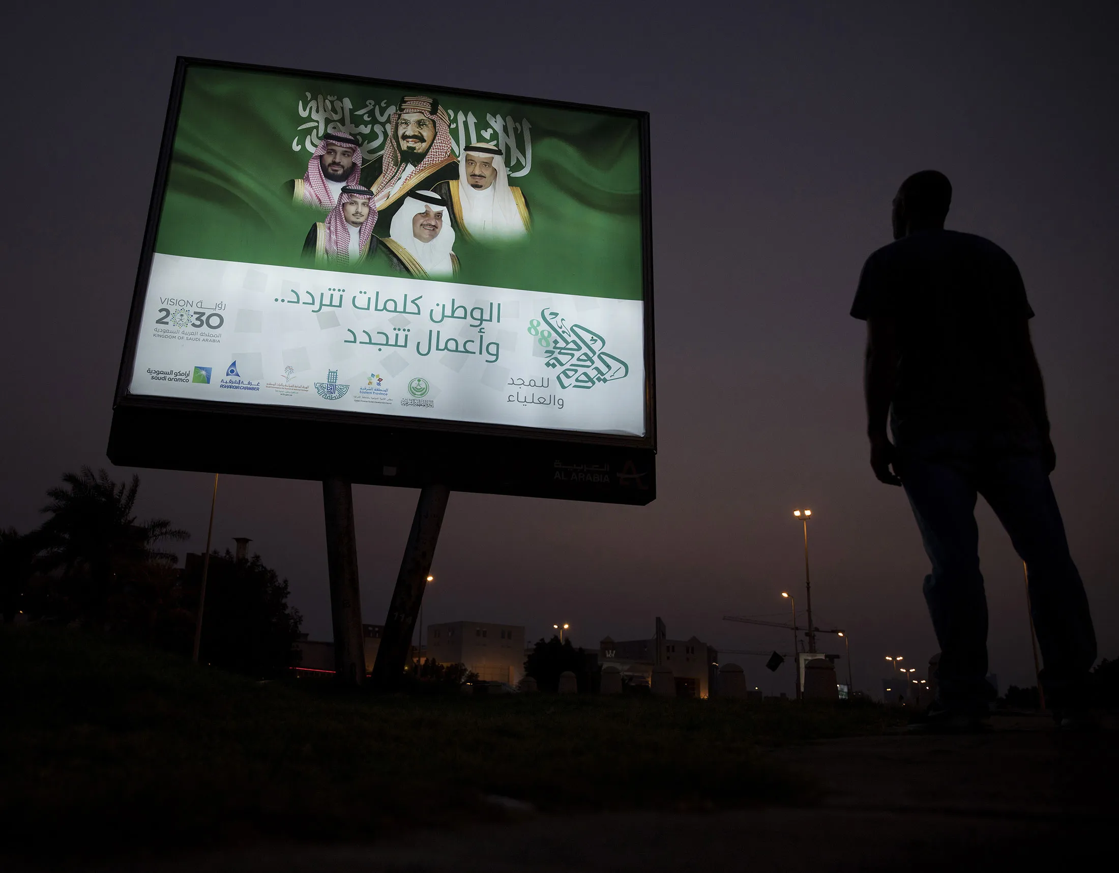 A pedestrian pauses near an illuminated sign showing portraits of the Saudi Arabian Royal family in Dhahran, Saudi Arabia