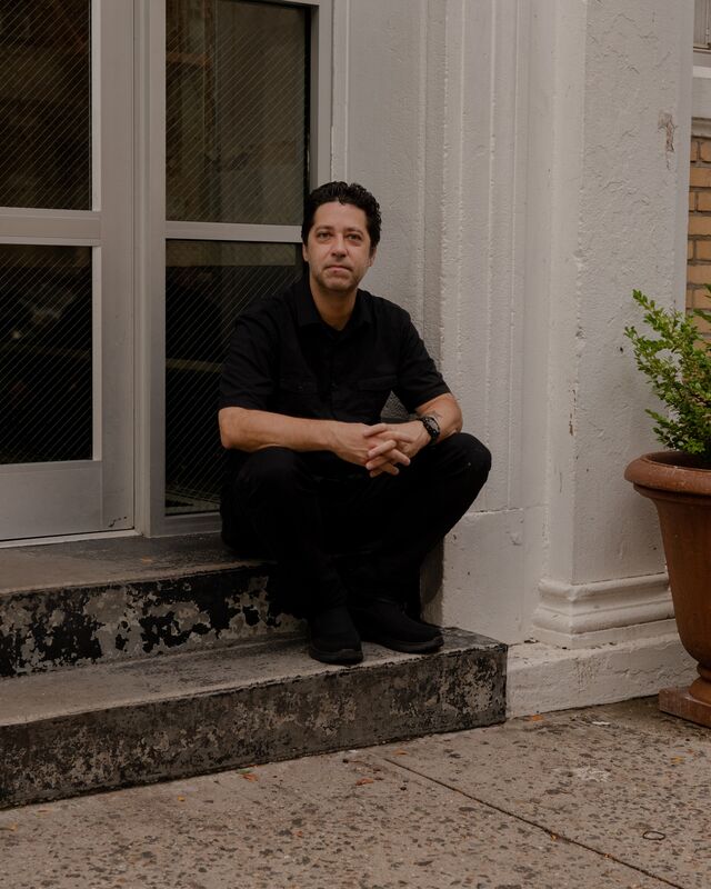 A middle-aged man, dressed in all black, sits with his hands clasped on a stoop.
