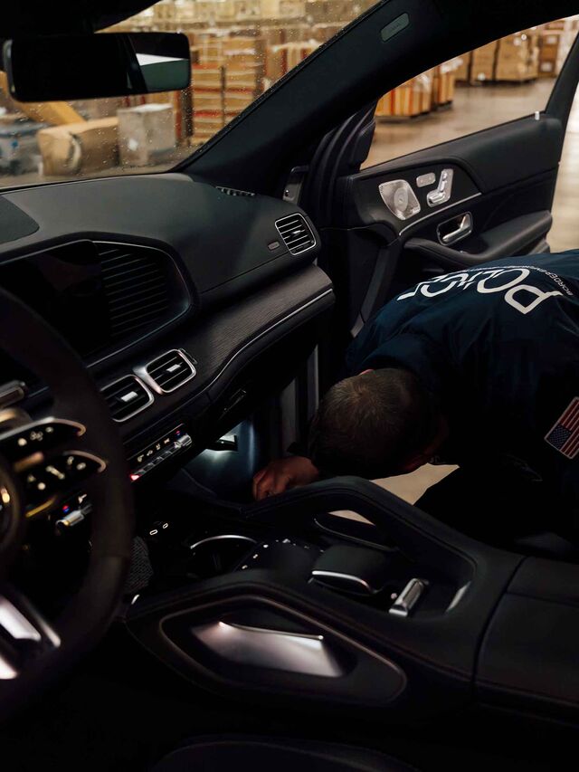 Interior photo showing a drive panel and front seats of a car; on the passenger side the back of a police offer is seen squatting with a flashlight, examining the lower portion of the vehicle.