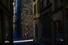 A woman walks in cobbled-stone street between stone buildings. It's a sunny morning but the sun is blocked by the buildings.