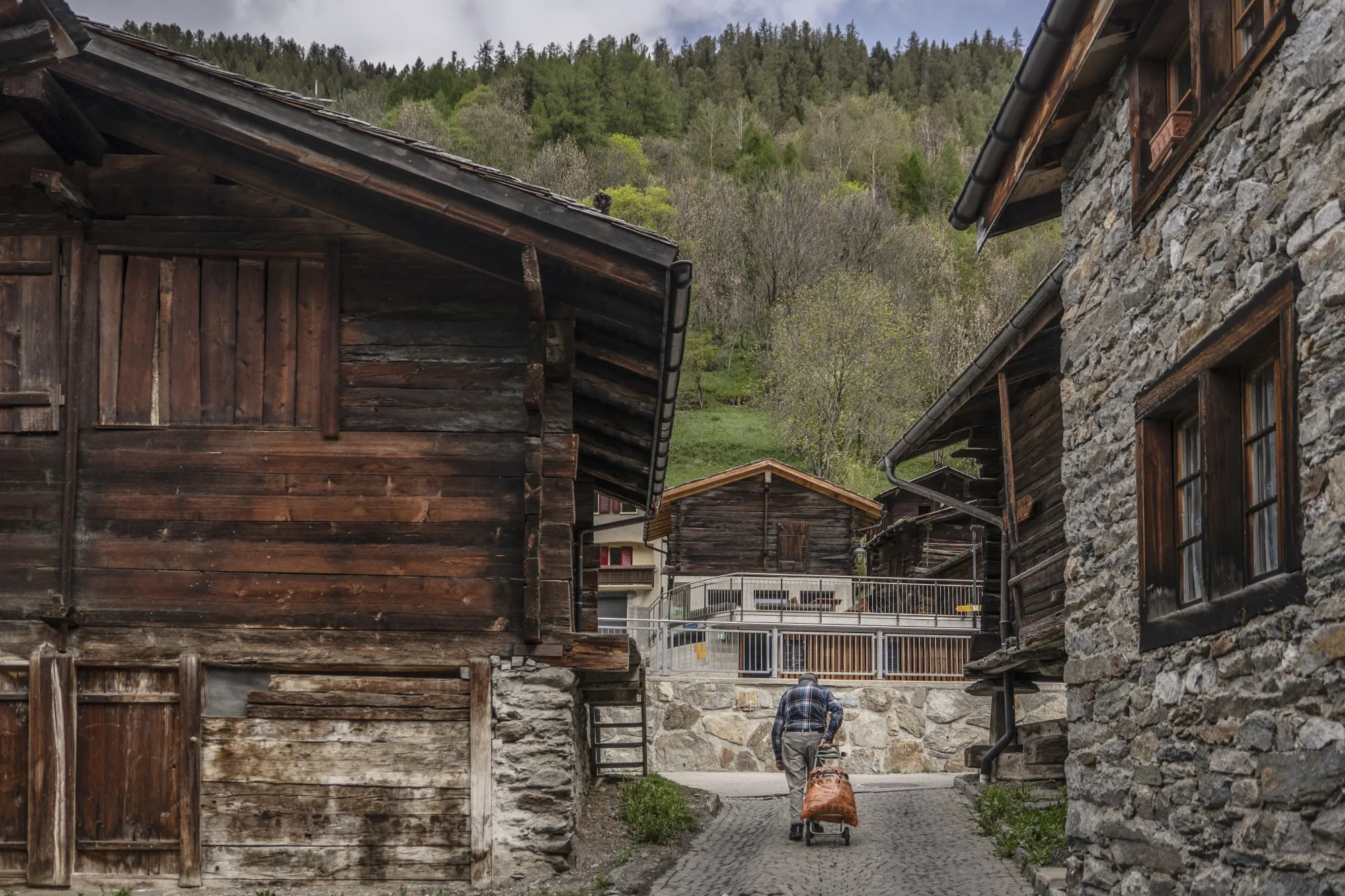 A man pulls a cart up a brick-lined road, in between two rustic buildings made of stone and weathered wood.