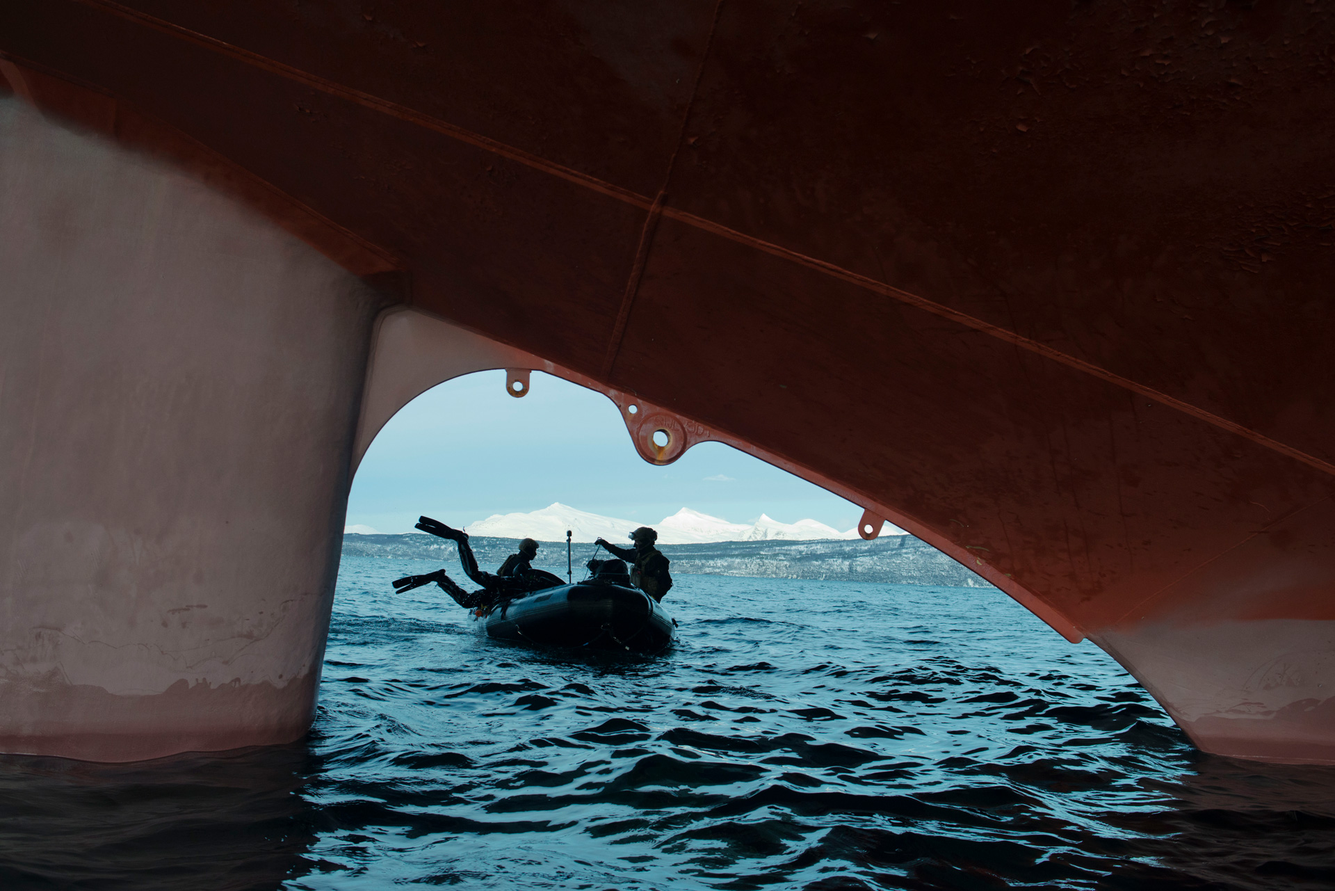 A small rubber boat with several people on it is glimpsed in silhouette through a gap in the red keel of a large ship floating in dark blue waters. One person can be seen wearing divers' flippers with their legs dangling over the side of the small boat. White peaks that may be snow-covered mountains or icebergs can be glimpsed in the far distance.