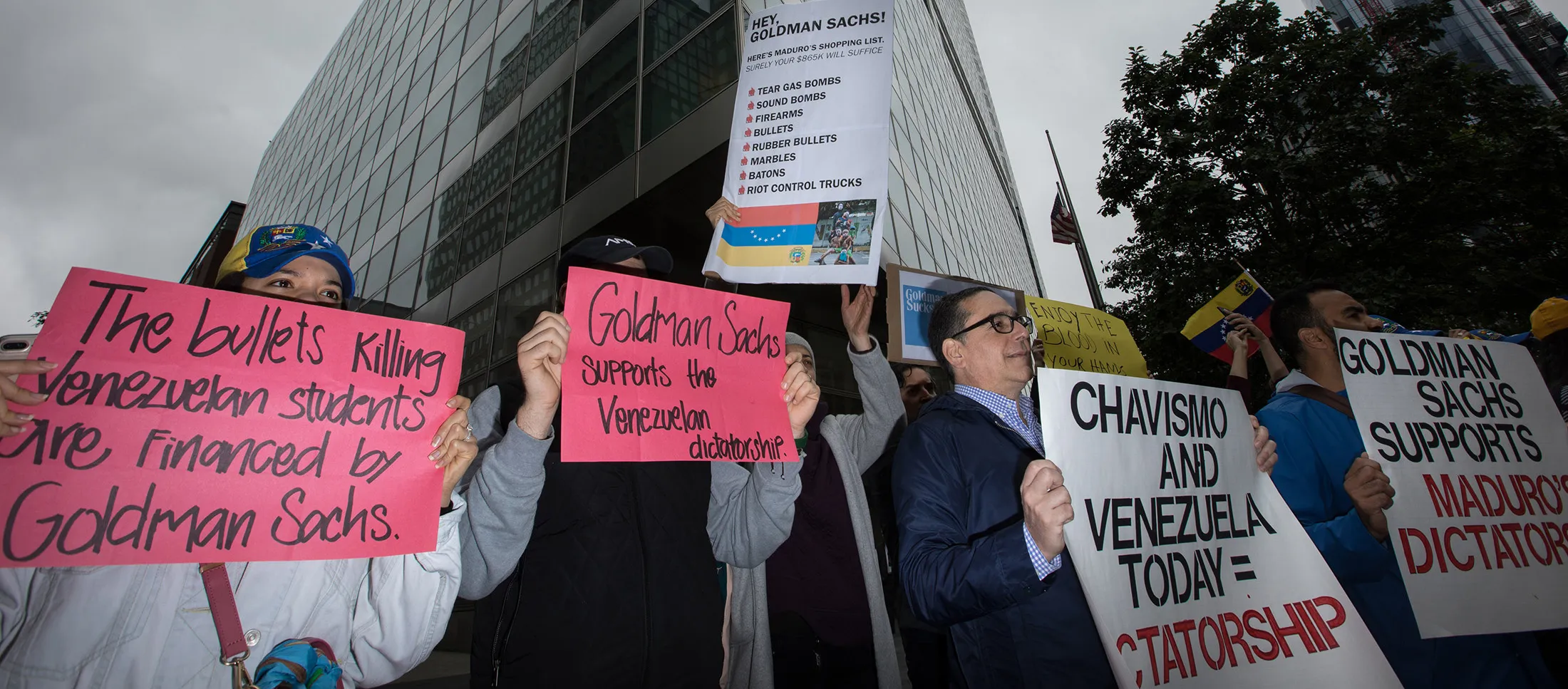 Demonstrators hold signs during a protest outside of the Goldman Sachs Group Inc. headquarters in New York, U.S., on Tuesday, May 30, 2017. Goldman faces a probe by Venezuela's opposition leaders after buying bonds issued in 2014 by the state oil company, a purchase some lawmakers said bolsters President Nicolas Maduro as he grapples with accusations of human-rights violations.
