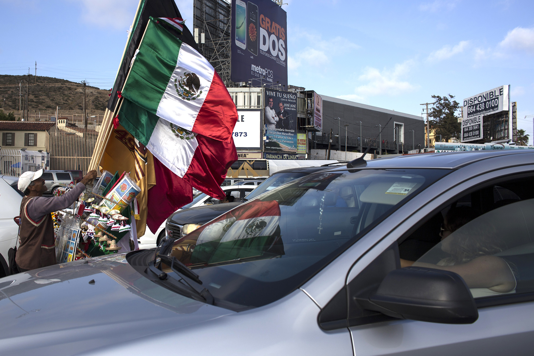 A street vendor sells flags and other wares as vehicles wait in line for re-entry into the United States of America on the Mexican side of the San Ysidro port of entry in Tijuana, Mexico, on Tuesday, June 7, 2016. The peso weakened, posting its biggest loss against the dollar on a closing basis in more than a month.
