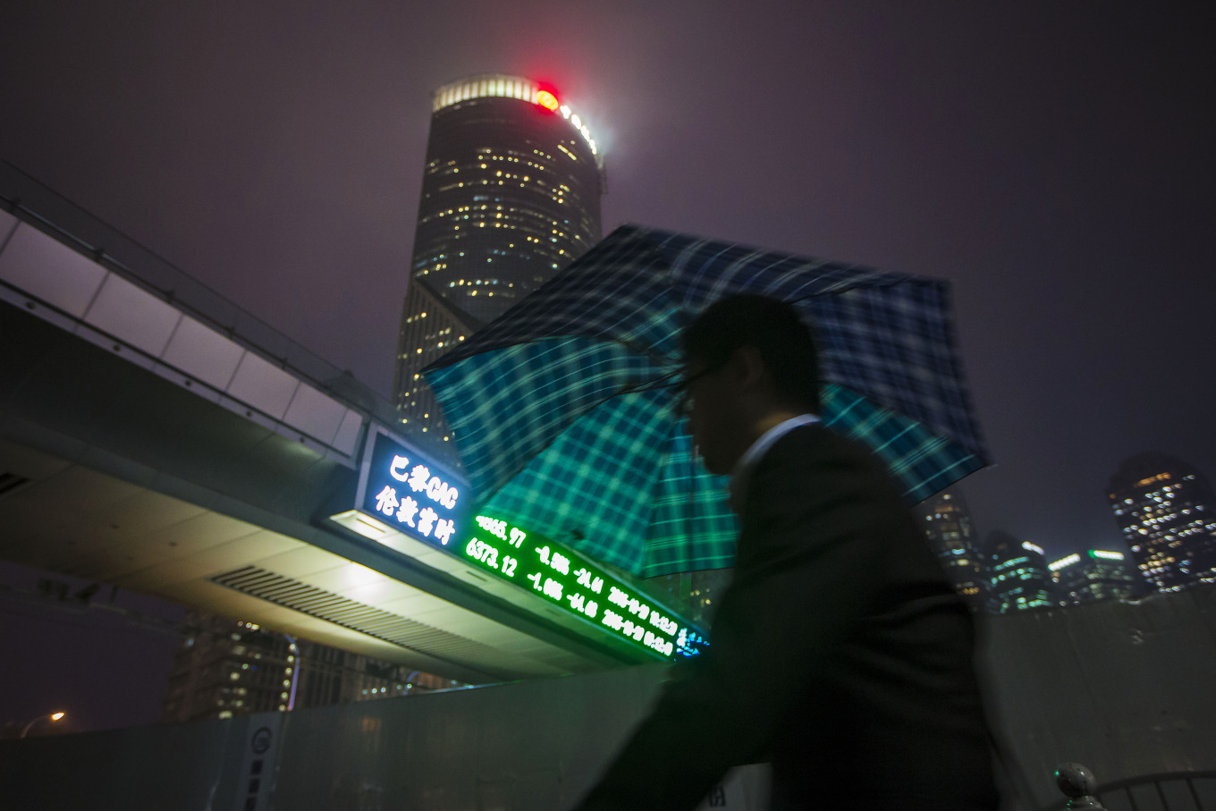 A pedestrian holding an umbrella walks past a pedestrian footbridge featuring an electronic stock ticker at night in the Lujiazui district of Shanghai, China.