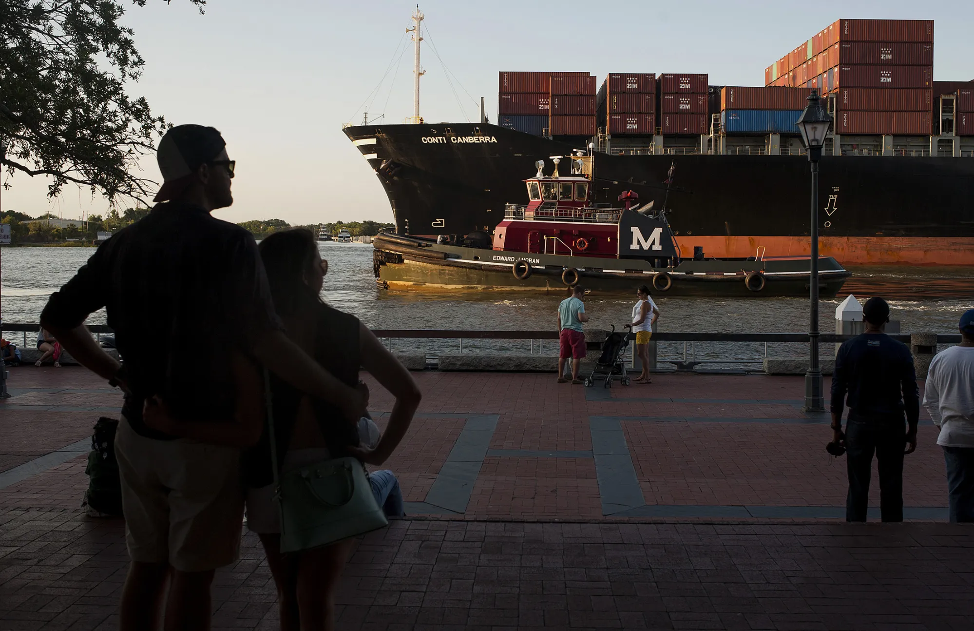 Pedestrians watch from River Street as a cargo ship and a tug boat travel into the Port of Savannah.