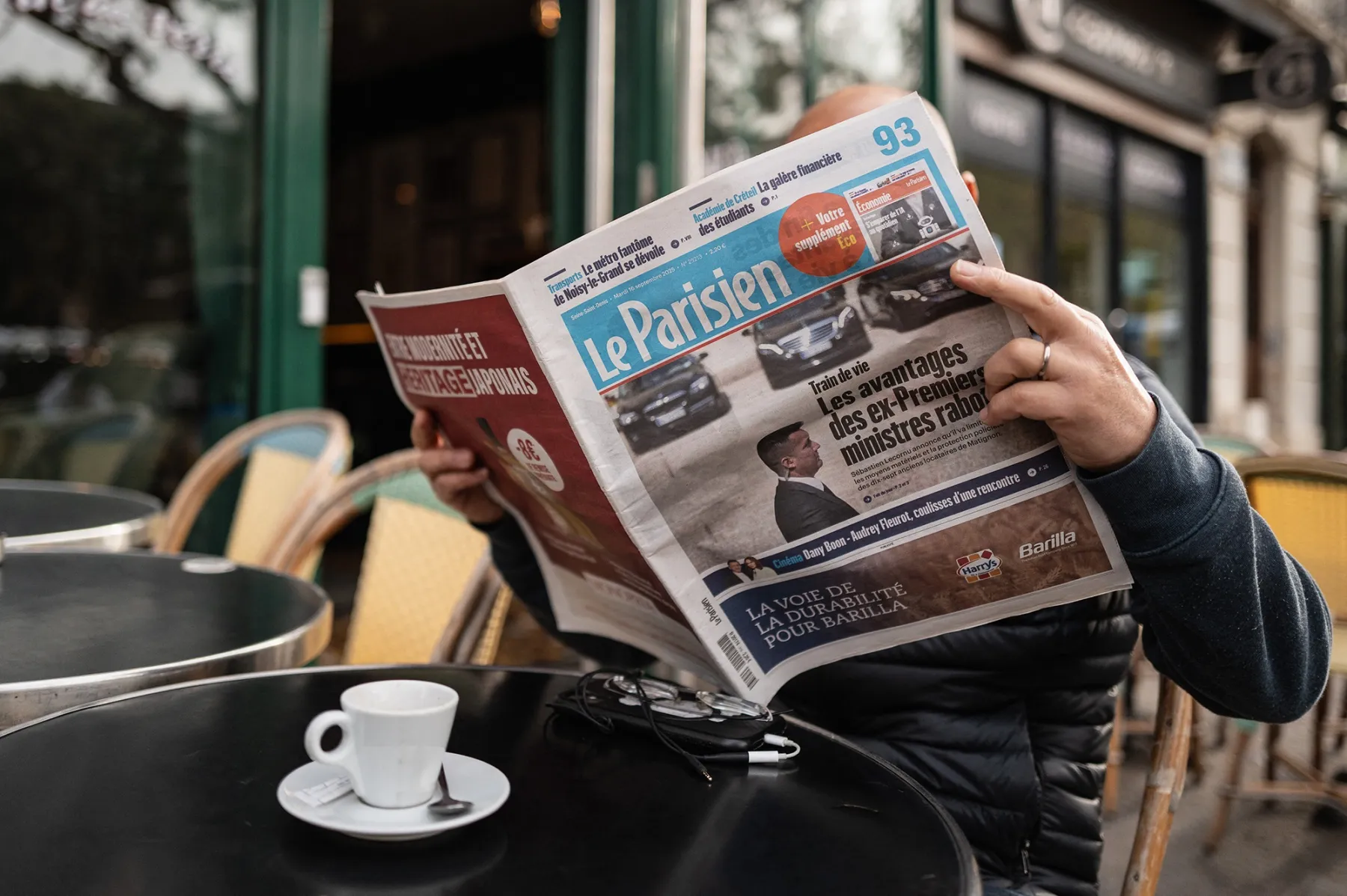 A man sits alone at a cafe table reading the newspaper Le Parisien.