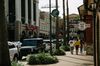 Wide-angle picture of a shopping district in Baton Rouge. It's daytime and bright, many cars are parked on the street. In the center of the picture is a large paved sidewalk lined up with vegetation and high trees (the sidewalk is large enough for three people to be walking side by side, their back turned to the photographer). Many store signs are visible, one reads Bistro bin 77 Wine, another towards the back reads Barnes and Nobles. The buildings are two stories high, except for one on the left of the picture that has large white colones in front of it and is more imposing. The sign in front of it reads "CineMark".