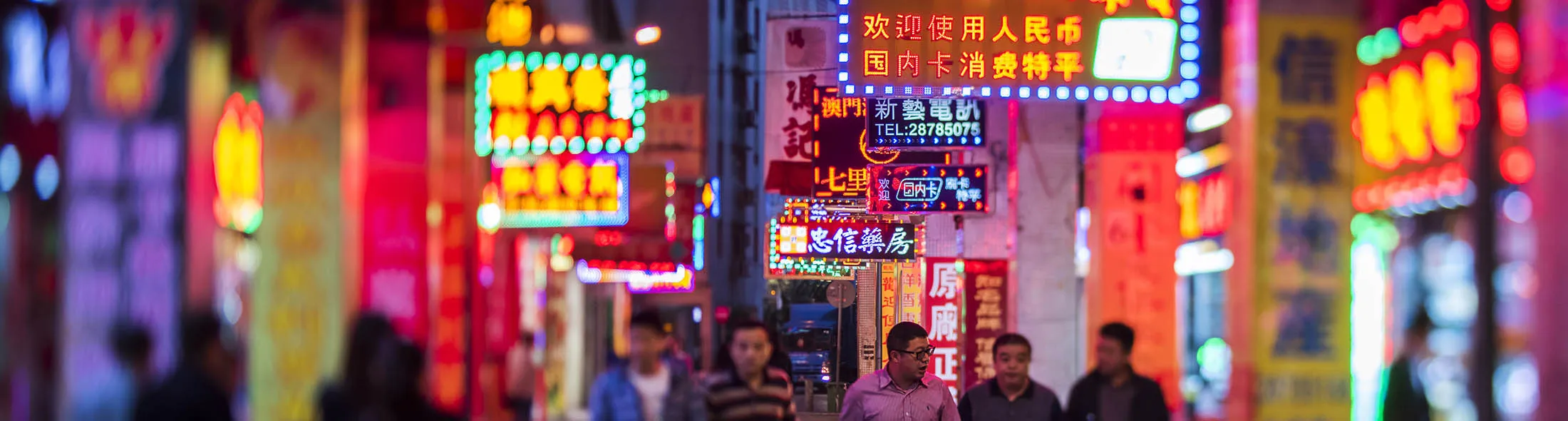 Pedestrians walk along a street with illuminated signs in this photograph taken with a tilt-shift lens at night in Macau, China, on Tuesday, Dec. 01, 2015. Macau's casino revenue fell for the 18th straight month in November, as China's moves to curb illicit money flows from the mainland deterred the high-stakes players who rely on junket promoters for betting loans.
