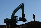 An excavator operates next to a worker at the construction site of a residential building