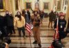 A topless man wearing fur hats and face paint stands in the middle of the hallway, holding an American flag, with other Trump supporters stand around