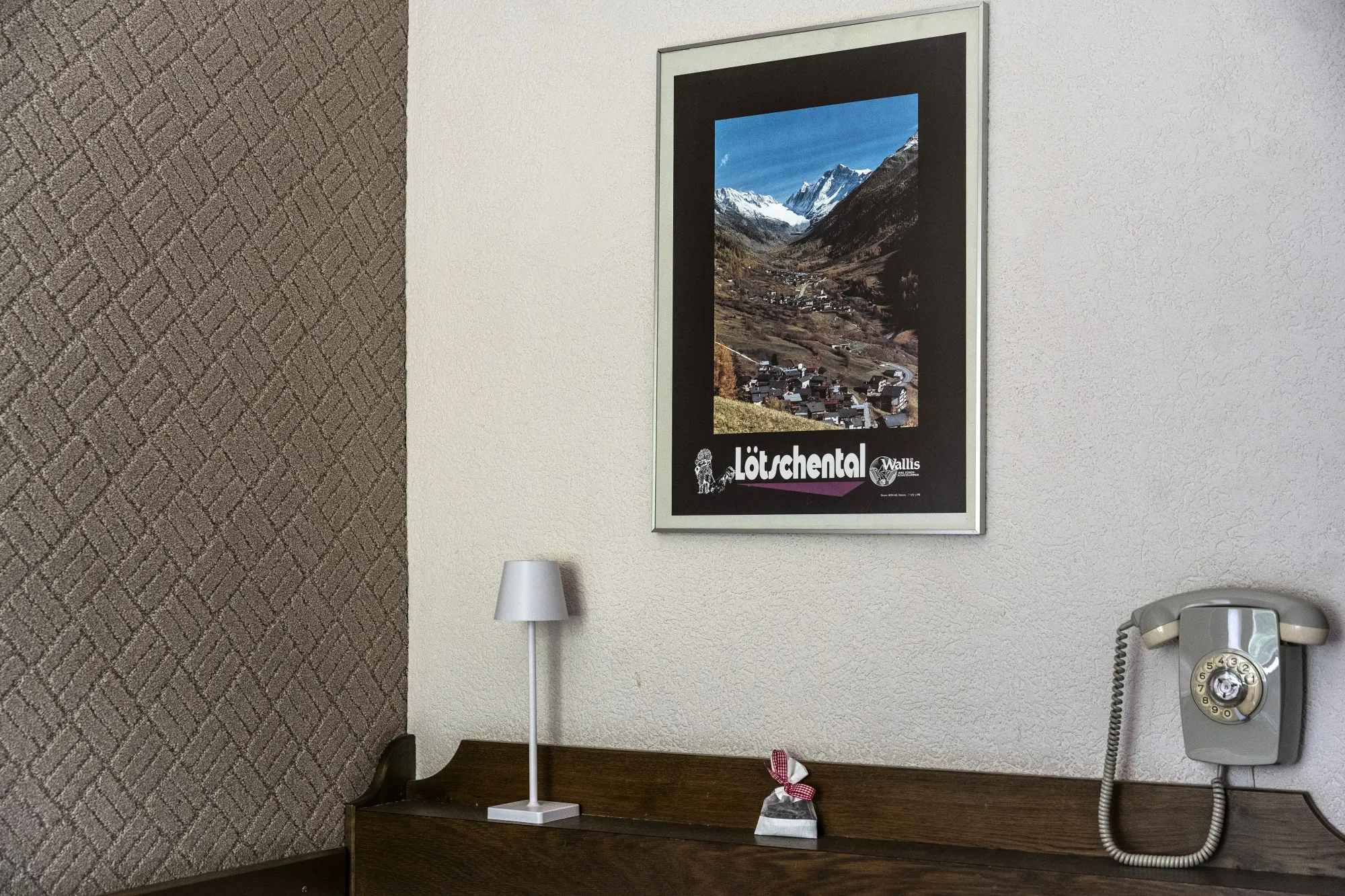 A framed poster hangs on a wall above a small table with a lamp and a telephone. The poster shows a scenic photo of the valley with homes in the foreground and snowcapped peaks in the background.