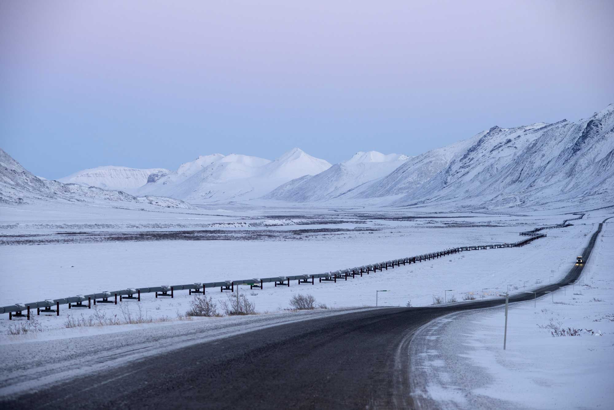 The Trans-Alaska Pipeline and the Dalton Highway, black lines against white snow, running parallel into icy mountains and lavender skies in the distance.