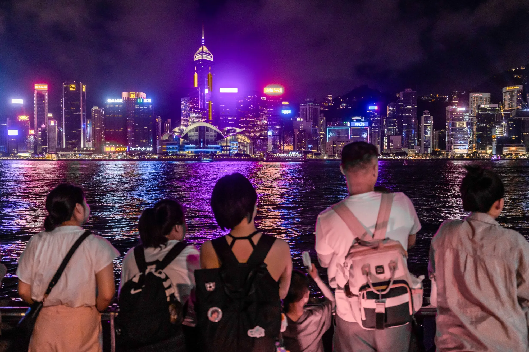 Pedestrians look out across Victoria Harbour towards the Hong Kong Island skyline in the Tsim Sha Tsui district of Hong Kong, China, on Sunday, June 23, 2024. 