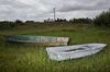 Two row boats, one with a faded green design and the other gray and weathered, sit on green grass with two houses uphill against an overcast sky.