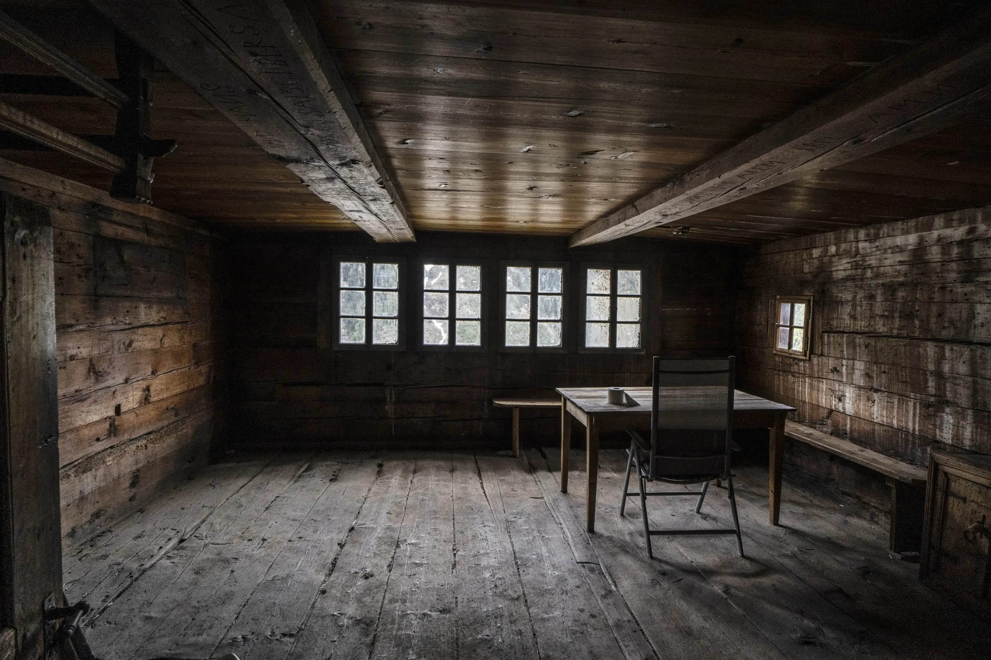 A plain wooden room, filled with light from windows against one wall, is empty except for one table and chair.