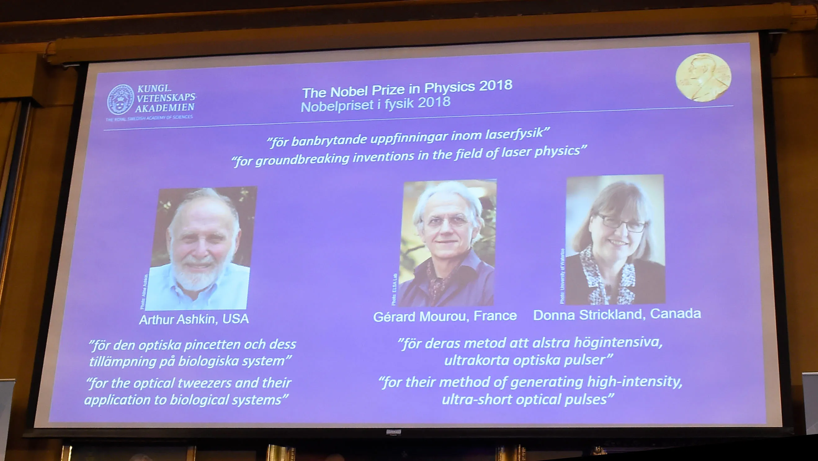 A screen displays portraits of Arthur Ashkin, Gerard Mourou  and Donna Strickland in Stockholm.