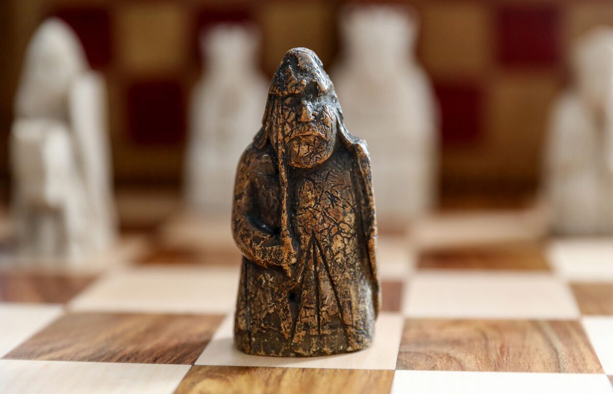  A brown chess piece, made of deer antler, carved with a human face and wearing a cloak, stands on a wooden chessboard with other chess pieces in the background.